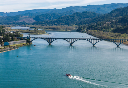 Aerial view of the Isaac Lee Patterson Bridge spanning the Rogue River with a boat leaving a wake in the water surrounded by lush green hills in Gold Beach Oregon United States.