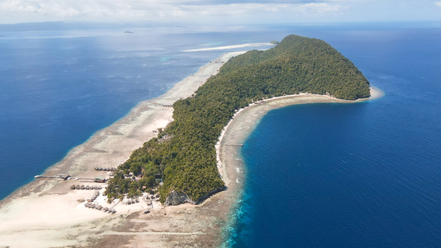 Aerial view of Raja Ampat island with lush vegetation white sand beaches and bungalows surrounded by turquoise reefs and deep blue ocean in Raja Ampat Southwest Papua Indonesia.