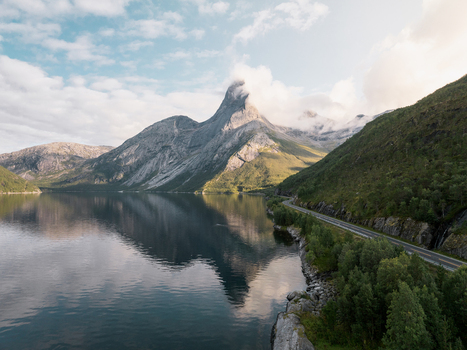 Aerial view of Stetind mountain reflecting in the calm fjord waters with a coastal road winding along the rocky shoreline in Narvik Municipality Nordland Norway. Aerial view of Stetind mountain reflecting in the calm fjord waters with a coastal road winding along the rocky shoreline in Narvik Municipality Nordland Norway.