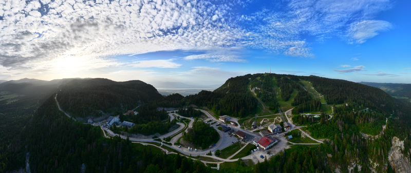 Aerial view of the Col de la Faucille mountain pass with winding roads dense pine forests and alpine resort buildings under a dramatic sunset sky in Gex Auvergne Rhone Alpes France.