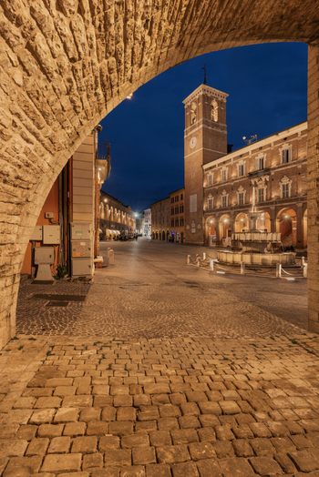 View of the historic Piazza del Comune in Fabriano featuring the illuminated Stura Fountain and the medieval Podesta Palace framed by a stone archway in Fabriano Marche Italy. View of the historic Piazza del Comune in Fabriano featuring the illuminated Stura Fountain and the medieval Podesta Palace framed by a stone archway in Fabriano Marche Italy.