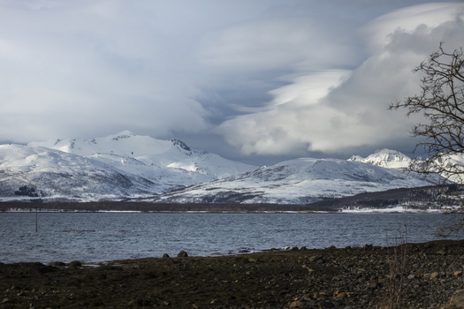 View of snow covered mountains across a calm fjord under a dramatic cloudy sky from a rocky shoreline in Tromso Troms Norway.