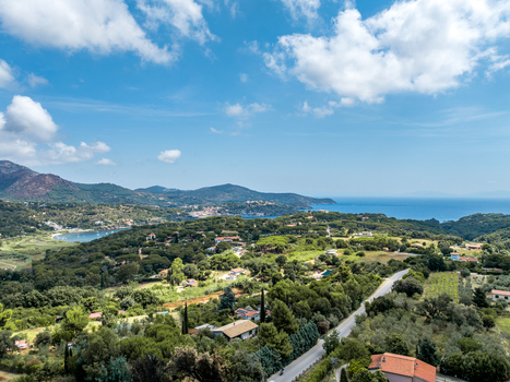 Aerial view of the coastal landscape with lush green hills winding roads scattered houses and the Mediterranean sea under a blue sky in Porto Azzurro Toscana Italy.