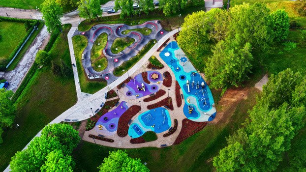 Aerial view of a modern playground with colorful rubber flooring and a winding pump track surrounded by lush green trees in Latvia. Aerial view of a modern playground with colorful rubber flooring and a winding pump track surrounded by lush green trees in Latvia.