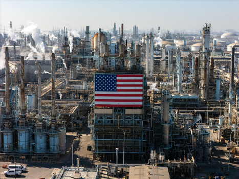 Aerial view of an oil refinery with a large American flag on a central structure surrounded by piping storage tanks and smoking chimneys in Carson California United States.