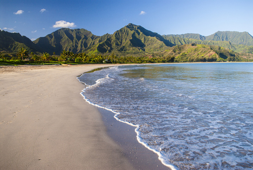 View of the sandy shore of Hanalei Bay with gentle waves and lush green mountains under a clear blue sky in Hanalei Hawaii United States. View of the sandy shore of Hanalei Bay with gentle waves and lush green mountains under a clear blue sky in Hanalei Hawaii United States.