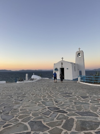 Porto Rafti Greece 21 September 2026 View of a small white Greek chapel by the sea at sunset with two people walking towards the entrance on a stone paved courtyard.