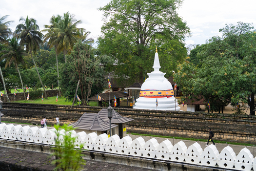 View of a white stupa surrounded by lush tropical trees stone walls and people walking within a temple complex in Kandy Central Province Sri Lanka. View of a white stupa surrounded by lush tropical trees stone walls and people walking within a temple complex in Kandy Central Province Sri Lanka.