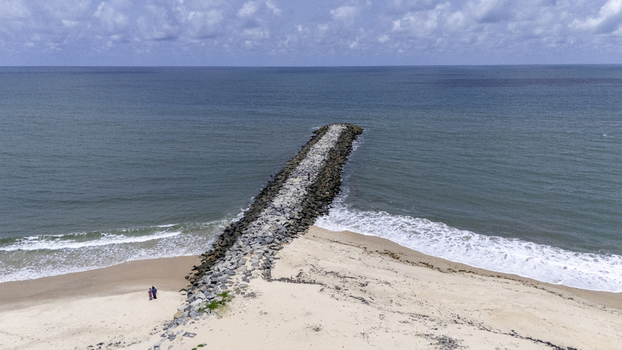Aerial view of a stone breakwater extending into the ocean from a sandy beach with people standing near the shore under a cloudy sky in Lekki Lagos Nigeria. Aerial view of a stone breakwater extending into the ocean from a sandy beach with people standing near the shore under a cloudy sky in Lekki Lagos Nigeria.