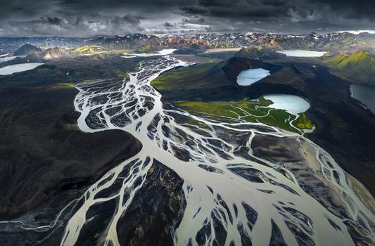 Aerial view of a vast braided glacial river system flowing through volcanic highlands with turquoise lakes and mossy mountains under a dramatic sky in Iceland.
