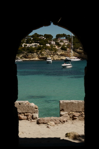 View of a turquoise bay with boats and rocky coastline framed by a stone archway in Mallorca Balearic Islands Spain.