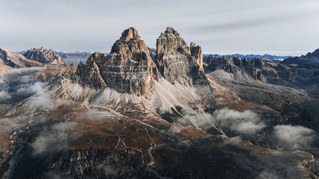 Aerial view of the Tre Cime di Lavaredo featuring jagged limestone peaks winding mountain paths and low clouds over the rocky alpine landscape Auronzo di Cadore Veneto Italy. Aerial view of the Tre Cime di Lavaredo featuring jagged limestone peaks winding mountain paths and low clouds over the rocky alpine landscape Auronzo di Cadore Veneto Italy.