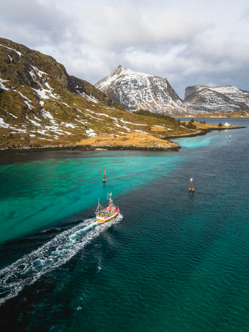 Aerial view of a fishing boat slicing through turquoise waters contrasting with the rugged snow dusted mountains of Kåkernbrua Ramberg Nordland Norway. Aerial view of a fishing boat slicing through turquoise waters contrasting with the rugged snow dusted mountains of Kåkernbrua Ramberg Nordland Norway.