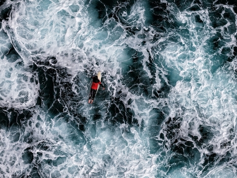 Aerial view of a surfer in a red shirt paddling through churning white foam and dark blue waves at Praia do Areal de Santa Barbara in Ribeira Grande Acores Portugal. Aerial view of a surfer in a red shirt paddling through churning white foam and dark blue waves at Praia do Areal de Santa Barbara in Ribeira Grande Acores Portugal.