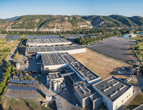 Aerial view of an industrial facility and warehouse complex integrated with solar photovoltaic farms set against rolling hills and a river in Le Pouzin Auvergne Rhone Alpes France.