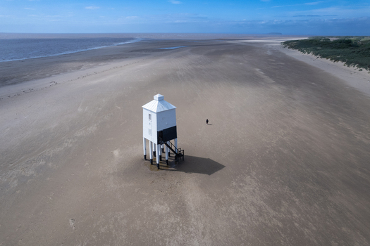 Aerial view of the Burnham on Sea Low Lighthouse standing on the vast sandy beach with a lone person walking nearby under a clear blue sky in Burnham on Sea England United Kingdom.