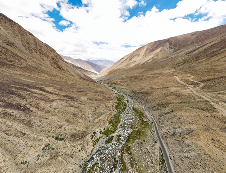 Aerial view of a winding road and a stream flowing through the arid mountain valley of the Himalayas under a bright blue sky with scattered clouds in Ladakh Division Ladakh India. Aerial view of a winding road and a stream flowing through the arid mountain valley of the Himalayas under a bright blue sky with scattered clouds in Ladakh Division Ladakh India.