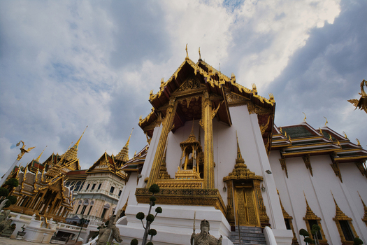 View of The Grand Palace featuring intricate gold ornamentation and traditional Thai architecture under a cloudy sky in Bangkok Thailand. View of The Grand Palace featuring intricate gold ornamentation and traditional Thai architecture under a cloudy sky in Bangkok Thailand.