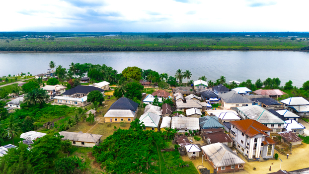 Aerial view of a riverside village featuring houses with metal roofs surrounded by lush green vegetation along a wide river in Minima Rivers Nigeria.