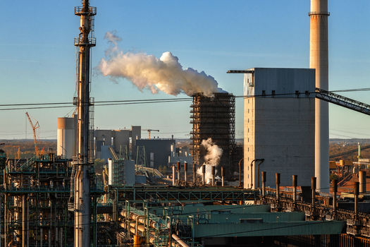 View of an industrial steel plant with smoking chimneys and complex piping under a clear blue sky during golden hour in Duisburg North Rhine Westphalia Germany.
