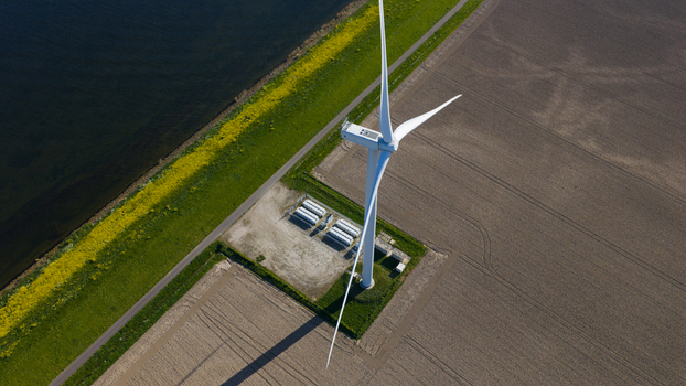 Aerial view of a large white wind turbine and battery storage units in a plowed field next to a water canal with yellow flowers in Ooltgensplaat Zuid Holland Netherlands. Aerial view of a large white wind turbine and battery storage units in a plowed field next to a water canal with yellow flowers in Ooltgensplaat Zuid Holland Netherlands.