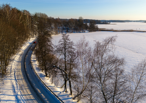 Aerial view of a red car driving on a snowy road winding through a winter forest next to a frozen lake under a clear blue sky Lithuania. Aerial view of a red car driving on a snowy road winding through a winter forest next to a frozen lake under a clear blue sky Lithuania.
