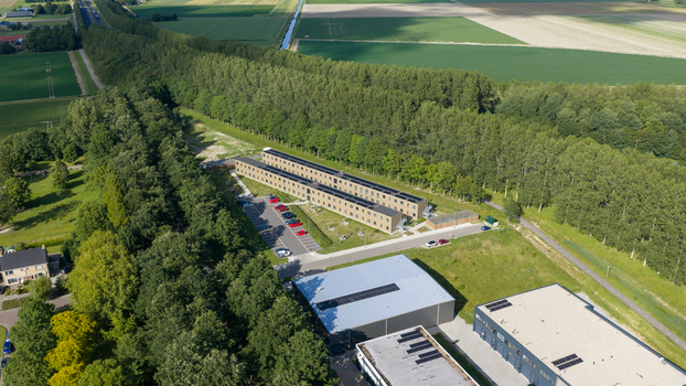 Aerial view of a modern building complex with solar panels adjacent parking lot with cars surrounded by trees and agricultural fields in Middenmeer North Holland Netherlands.