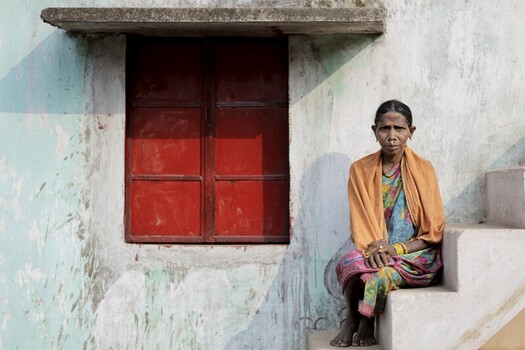 Odisha India 26 March 2026 View of a woman sitting on weathered steps beside a vibrant red window her orange shawl a splash of warmth against the cool textured wall. Odisha India 26 March 2026 View of a woman sitting on weathered steps beside a vibrant red window her orange shawl a splash of warmth against the cool textured wall.