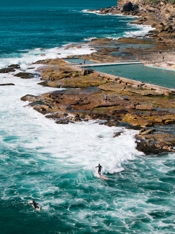 Aerial view of the rock pool surrounded by rocky coastline with surfers riding waves in the turquoise ocean water Dee Why New South Wales Australia. Aerial view of the rock pool surrounded by rocky coastline with surfers riding waves in the turquoise ocean water Dee Why New South Wales Australia.