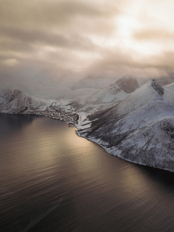 Aerial view of a serene dark fjord mirroring the overcast sky contrasting with the snow dusted mountains of the Norwegian landscape Fjordgård Troms Norway. Aerial view of a serene dark fjord mirroring the overcast sky contrasting with the snow dusted mountains of the Norwegian landscape Fjordgård Troms Norway.
