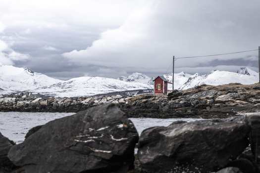 View of a small red wooden hut on a rocky shoreline with snow capped mountains under an overcast sky in Tromso Troms Norway.
