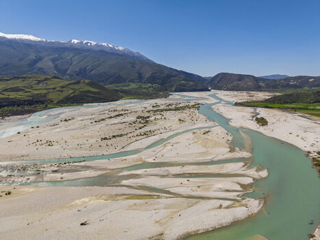 Aerial view of Vjosa Wild River National Park with its characteristic braided river channels gravel bars and snow capped mountains under a clear blue sky in Permet Gjirokaster County Albania. Aerial view of Vjosa Wild River National Park with its characteristic braided river channels gravel bars and snow capped mountains under a clear blue sky in Permet Gjirokaster County Albania.