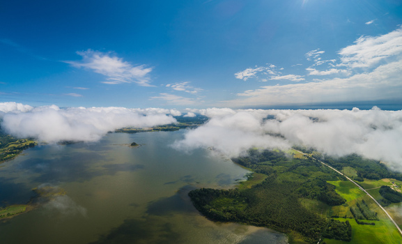 Aerial view of a serene lake surrounded by lush green forests fields and scattered clouds under a bright blue sky in Latvia. Aerial view of a serene lake surrounded by lush green forests fields and scattered clouds under a bright blue sky in Latvia.