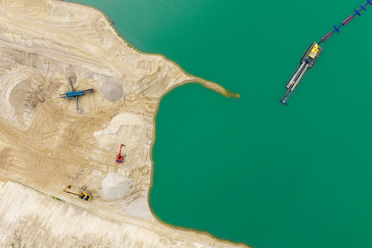 Aerial view of a sand and gravel mine with heavy machinery excavators and a floating dredge operating in a vibrant turquoise water pit in Masovian Voivodeship Poland.