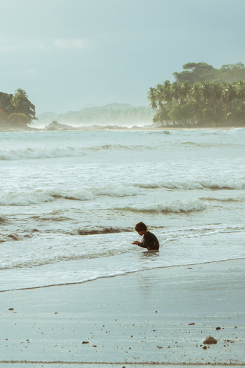 View of a young child playing in the shallow surf on a sandy beach with lush palm trees and ocean waves in Costa Rica.