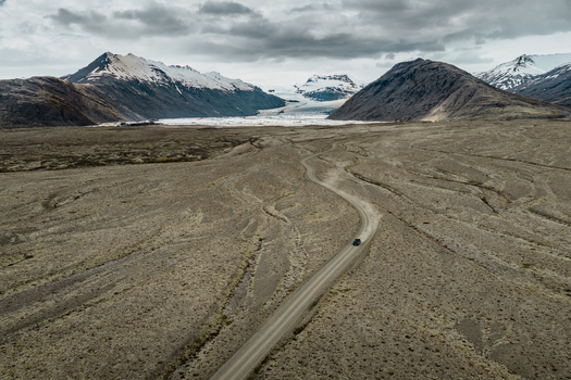 Aerial view of a winding gravel road leading towards the Vatnajokull glacier in a vast volcanic landscape under a cloudy sky in Iceland.