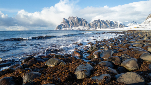 View of the rocky coastline at Uttakleiv Beach with snow capped mountains rising above the sea under a bright sky in Leknes Lofoten Norway.