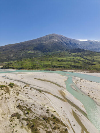Aerial view of Vjosa Wild River National Park featuring turquoise river channels expansive gravel riverbeds and lush green hills under a clear blue sky in Permet Gjirokaster County Albania. Aerial view of Vjosa Wild River National Park featuring turquoise river channels expansive gravel riverbeds and lush green hills under a clear blue sky in Permet Gjirokaster County Albania.
