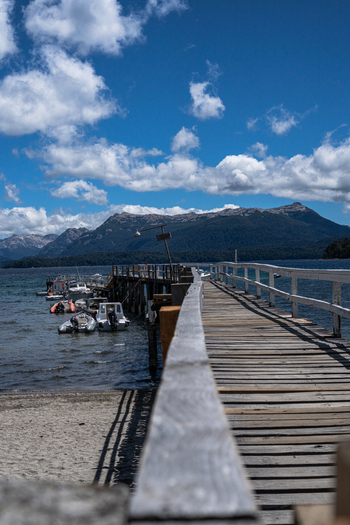View of Bahia Brava with a wooden pier extending into the lake small boats moored alongside and mountains under a blue sky in Villa La Angostura Neuquen Province Argentina. View of Bahia Brava with a wooden pier extending into the lake small boats moored alongside and mountains under a blue sky in Villa La Angostura Neuquen Province Argentina.