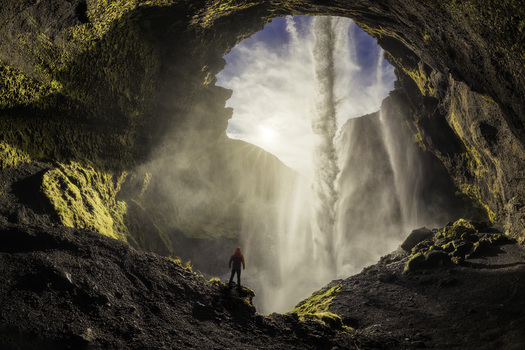 View of Kvernufoss waterfall from inside a mossy cave with a person in a red jacket standing on a ledge amidst mist and sunlight in Evindarholar Rangarthing eystra Iceland.
