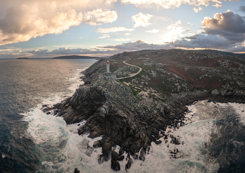 Aerial view of Faro de Punta Nariga on rocky cliffs with crashing waves and wind turbines under a cloudy sunset sky in Malpica A Coruna Spain. Aerial view of Faro de Punta Nariga on rocky cliffs with crashing waves and wind turbines under a cloudy sunset sky in Malpica A Coruna Spain.