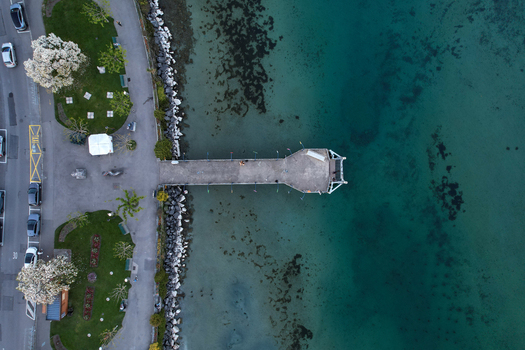 Aerial view of a concrete pier extending into the turquoise waters of Lake Geneva with blooming trees and a green park in Morges Vaud Switzerland. Aerial view of a concrete pier extending into the turquoise waters of Lake Geneva with blooming trees and a green park in Morges Vaud Switzerland.