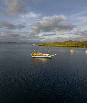 Aerial view of a traditional wooden phinisi boat anchored in calm blue waters near the lush coastline of Komodo Island with rolling hills in Komodo East Nusa Tenggara Indonesia.