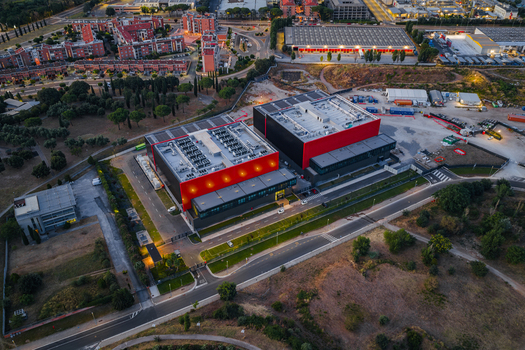 Aerial view of a modern datacenter facility featuring red and black exterior walls and rooftop cooling systems situated near roads and urban infrastructure in Settecamini Lazio Italy.