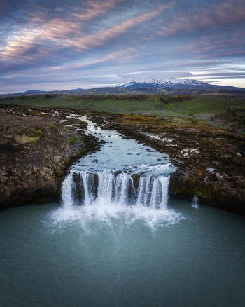 Aerial view of THjofafoss waterfall with turquoise water rugged volcanic terrain and a snow capped mountain under a dramatic sunset sky in Rangarthing ytra Iceland. Aerial view of THjofafoss waterfall with turquoise water rugged volcanic terrain and a snow capped mountain under a dramatic sunset sky in Rangarthing ytra Iceland.