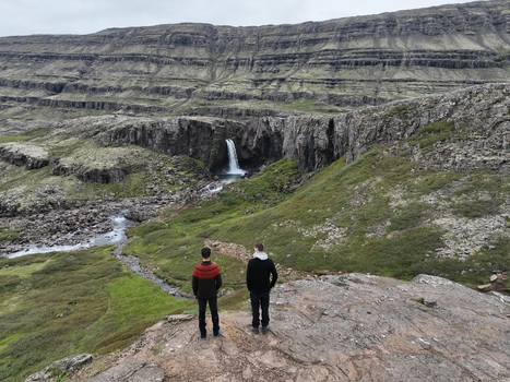 Aerial view of two people standing on a rocky cliff overlooking a waterfall cascading down a rugged mountain landscape with green moss and rocky terrain in Seydisfjordur Mulathing Iceland.
