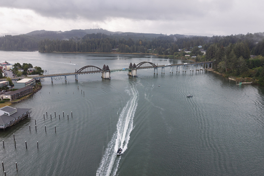 Aerial view of the historic Siuslaw River Bridge spanning the river with a motorboat leaving a white wake in the water surrounded by dense forested hills in Florence Oregon United States.