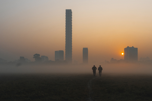 View of two figures walking through a misty field towards a modern skyline silhouetted against the rising sun Kolkata West Bengal India. View of two figures walking through a misty field towards a modern skyline silhouetted against the rising sun Kolkata West Bengal India.