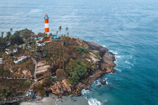 Aerial view of the iconic red and white Vizhinjam Lighthouse situated on a rocky cliff surrounded by lush palm trees and the blue waters of the Arabian Sea in Kovalam Keralam India. Aerial view of the iconic red and white Vizhinjam Lighthouse situated on a rocky cliff surrounded by lush palm trees and the blue waters of the Arabian Sea in Kovalam Keralam India.