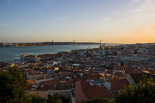 View of the 25 de Abril Bridge and the historic Lisbon skyline with red rooftops over the Tagus River during a golden sunset Lisbon Lisbon Portugal. View of the 25 de Abril Bridge and the historic Lisbon skyline with red rooftops over the Tagus River during a golden sunset Lisbon Lisbon Portugal.